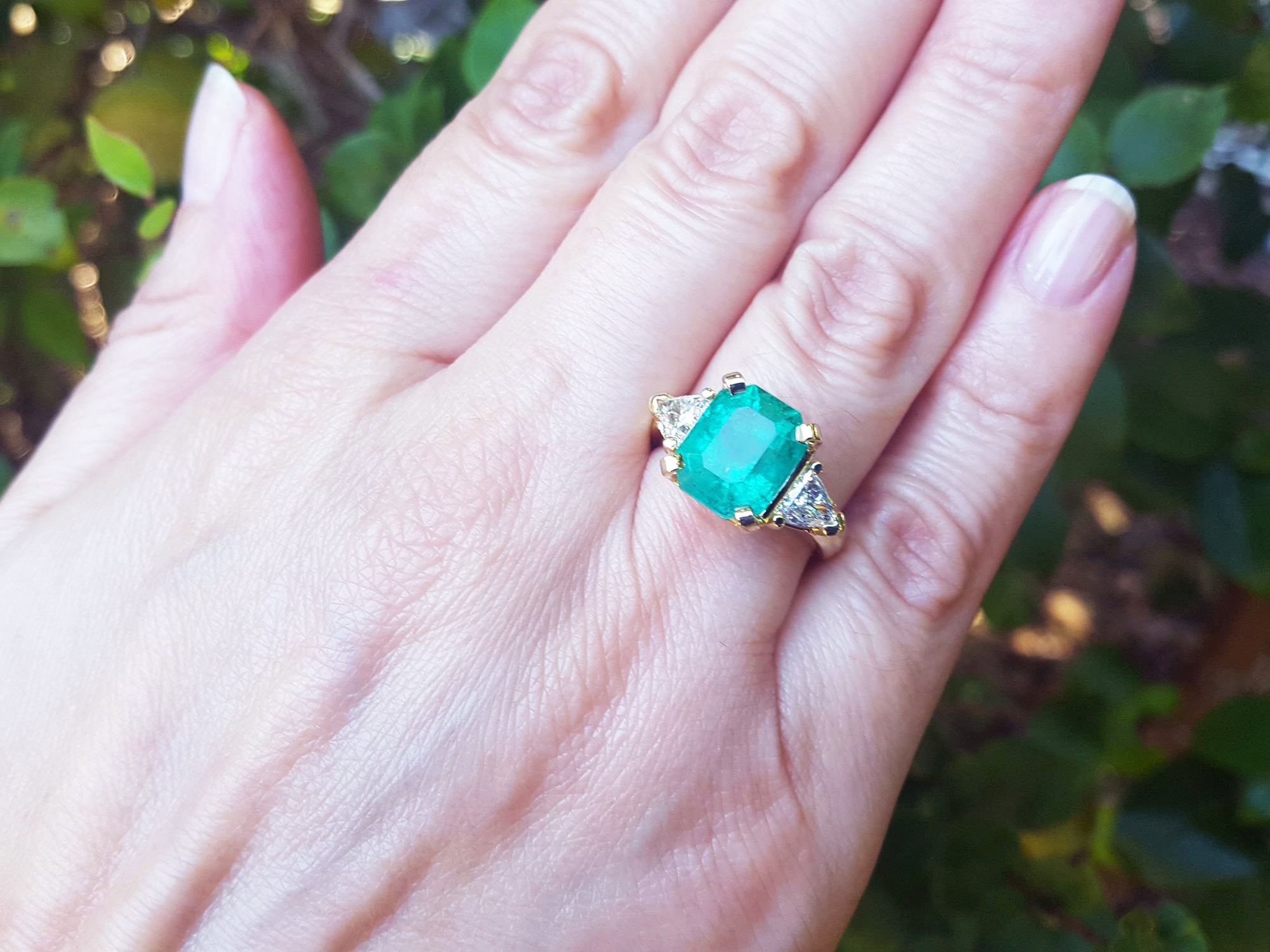 Hand wearing a Muzo Colombian emerald ring against a blurred natural background.