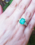 Hand wearing a Muzo Colombian emerald ring against a blurred natural background.