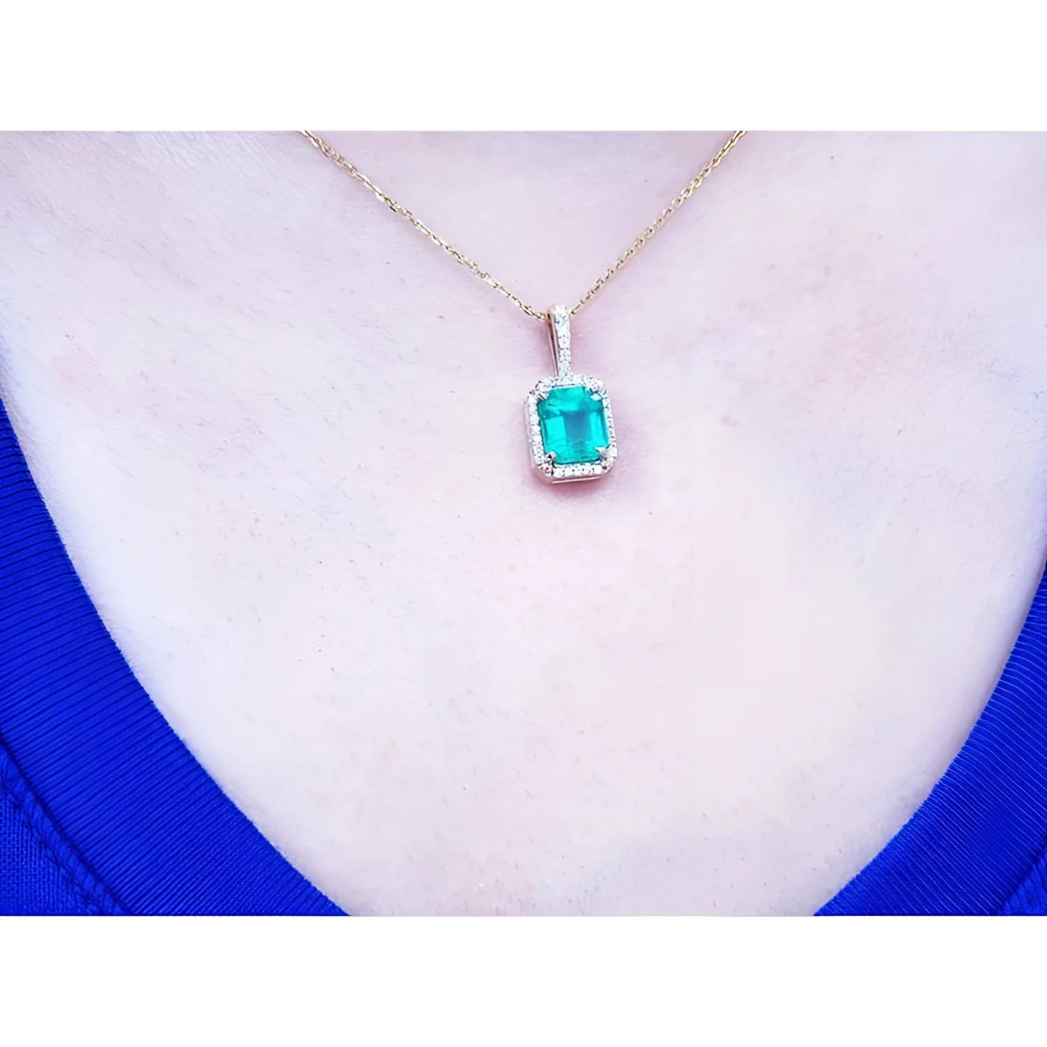 Emerald-cut Colombian emerald pendant with diamond halo on a 14k yellow gold chain, worn on a neck.