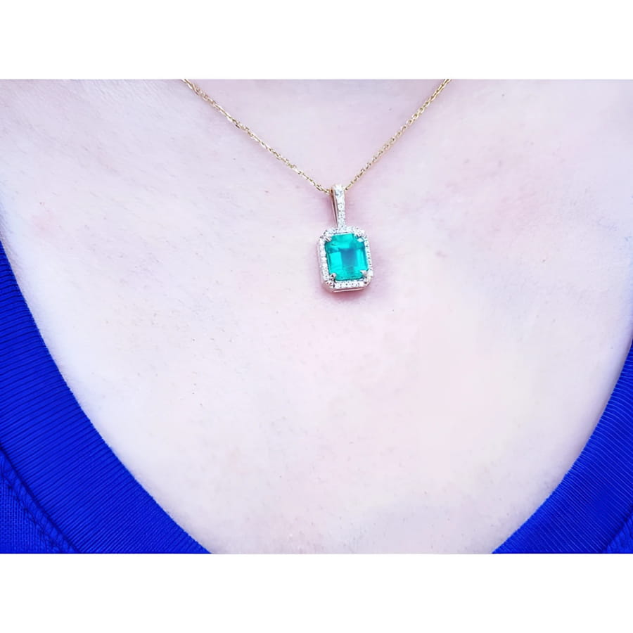 Emerald-cut Colombian emerald pendant with diamond halo on a 14k yellow gold chain, worn on a neck.