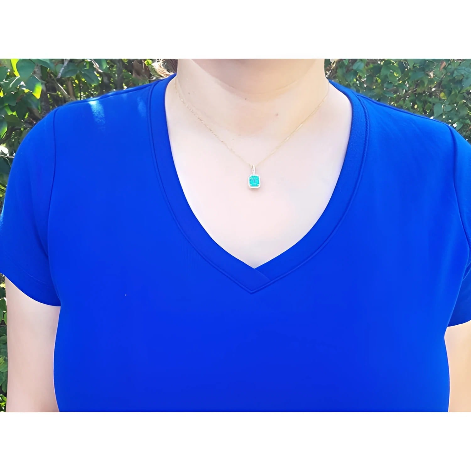 Emerald-cut emerald pendant with diamond halo on a thin gold chain, worn on a blue shirt.