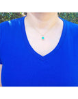 A woman wearing a blue shirt with a necklace featuring a halo diamond emerald pendant against a blurred green background.