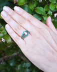 Hand wearing an enhancer emerald ring with a center diamond against a green leafy background.