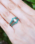 Hand wearing a ring with a pear-shaped center diamond and round emeralds gemstones against a blurred green background.