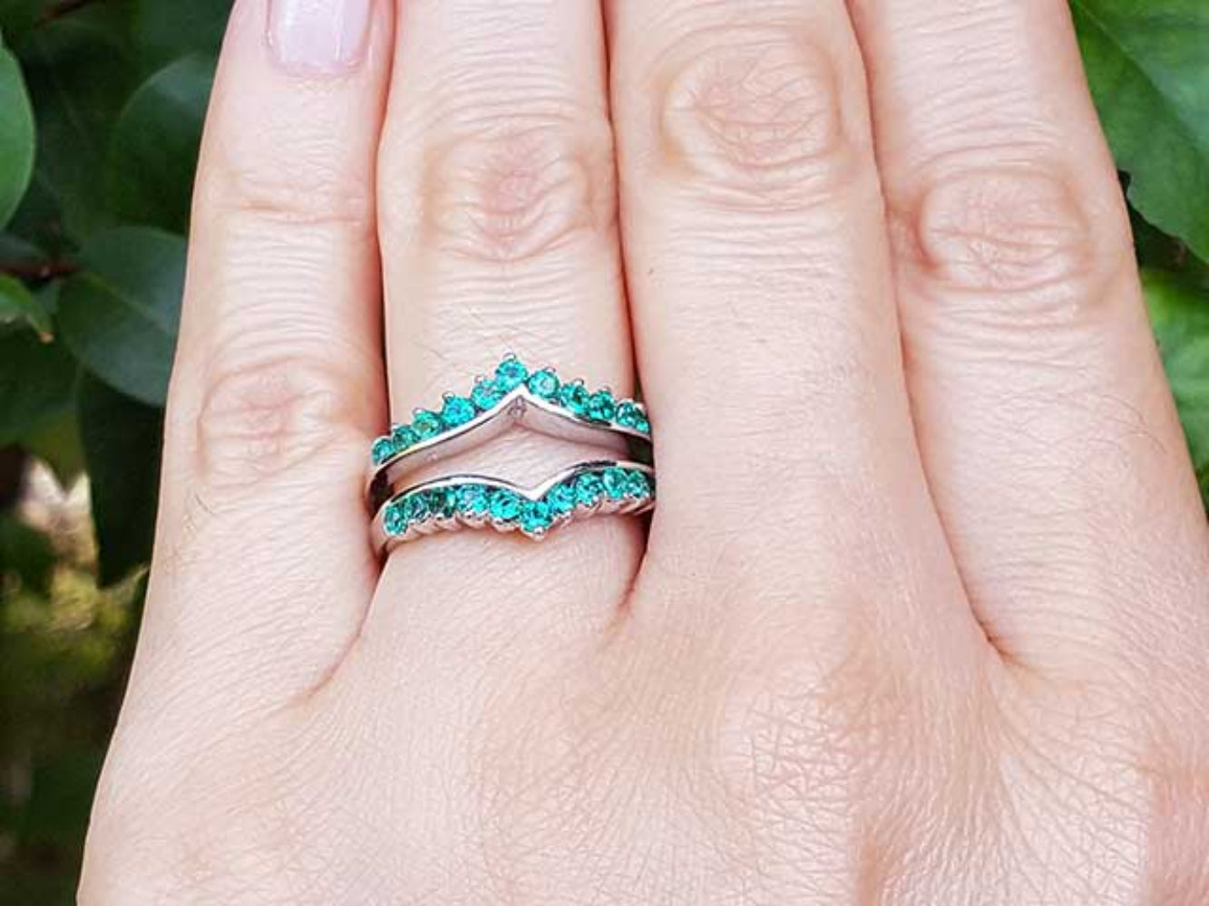 Close-up of a hand wearing a natural emerald enhancer ring with a blurred green leafy background.