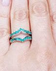 Close-up of a hand wearing a natural emerald enhancer ring with a blurred green leafy background.