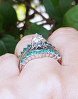 A white gold enhancer ring with emerald stones on a hand against a green leafy background.