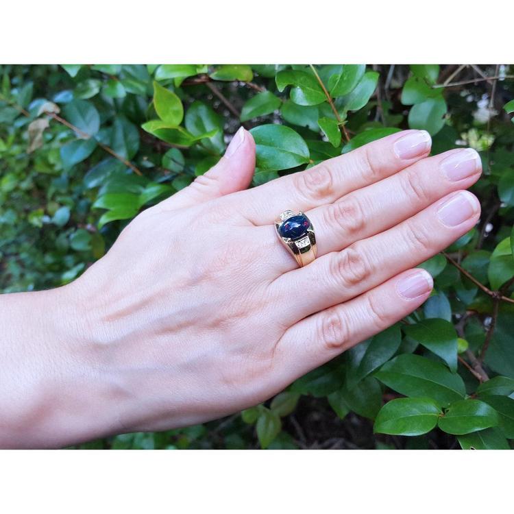 Hand wearing a ring with a black opal against a green leafy background.