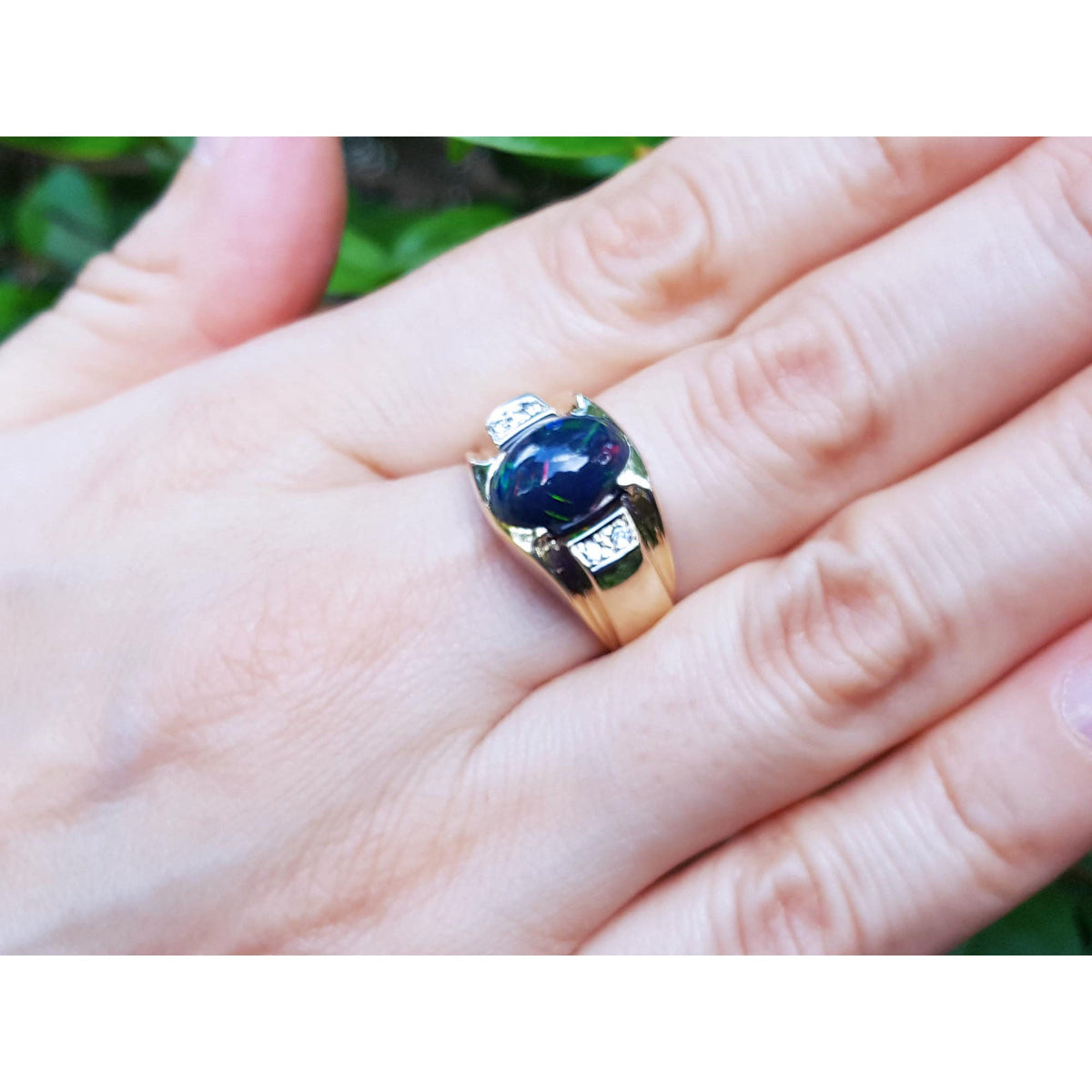 Gold ring with a black opal on a hand against a blurred green background.