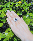 Hand wearing a silver ring for men with a red Ethiopian black opal against a green leafy background