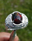 Silver ring for men with a red Ethiopian opal held by a hand against a blurred green background