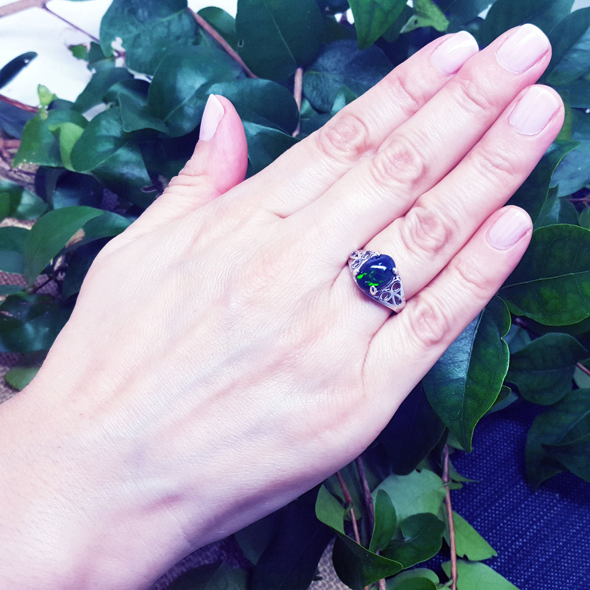 Hand wearing a sterling silver ring with a black Ethiopian black opal against a green leafy background.