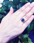 Hand wearing a sterling silver ring with a black Ethiopian black opal against a green leafy background.