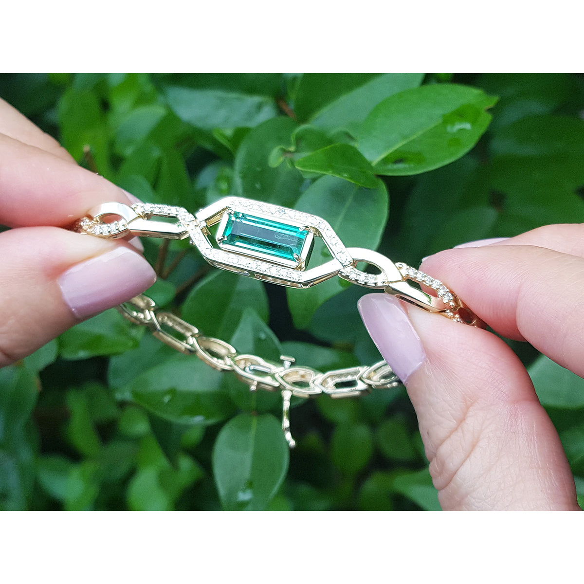 Gold bracelet with a Muzo colombian emerald held by a hand against a green leafy background