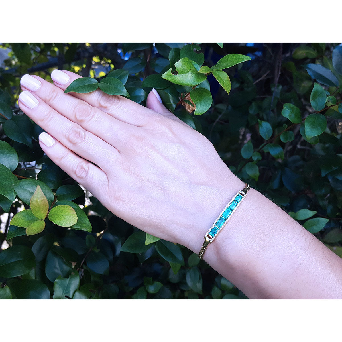 Hand wearing a five stone emeralds and diamonds bracelet with a green leafy background.