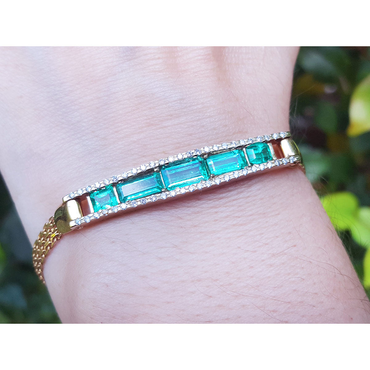 Gold bracelet with five natural emeralds on a wrist against a blurred natural background.