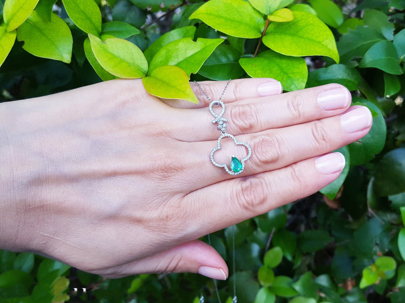 Hand displaying a four-leaf pendant with an emerald gemstone against a green leafy background.