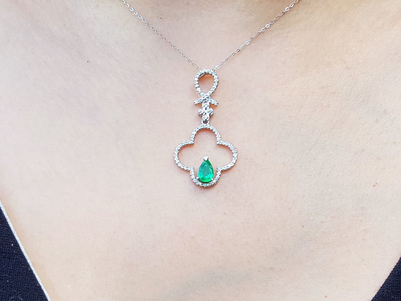 A white gold necklace with an emerald gemstone pendant on a person wearing a black top.