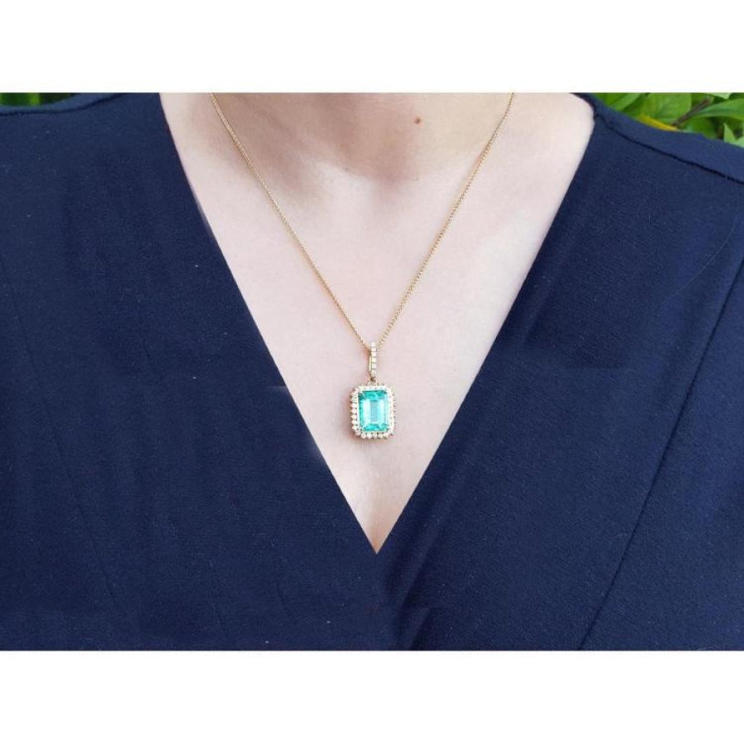 Emerald-cut emerald pendant with halo diamonds on an 18K gold chain, worn on a dark blue top.