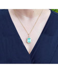 Emerald-cut emerald pendant with halo diamonds on an 18K gold chain, worn on a dark blue top.