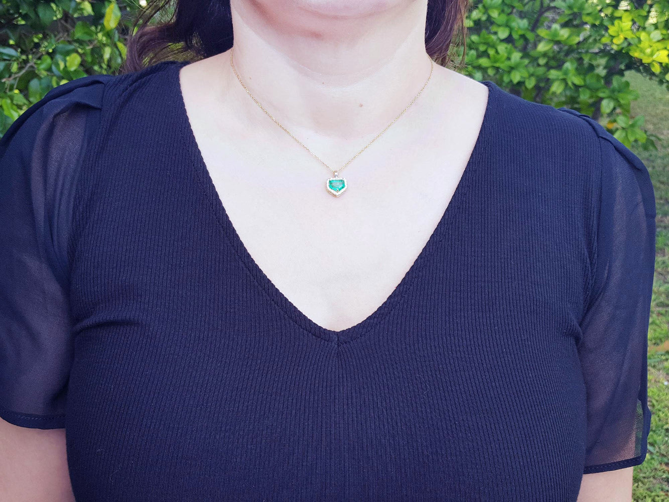 A woman wearing a dark blue top with a necklace featuring an emerald pendant against a blurred green background.