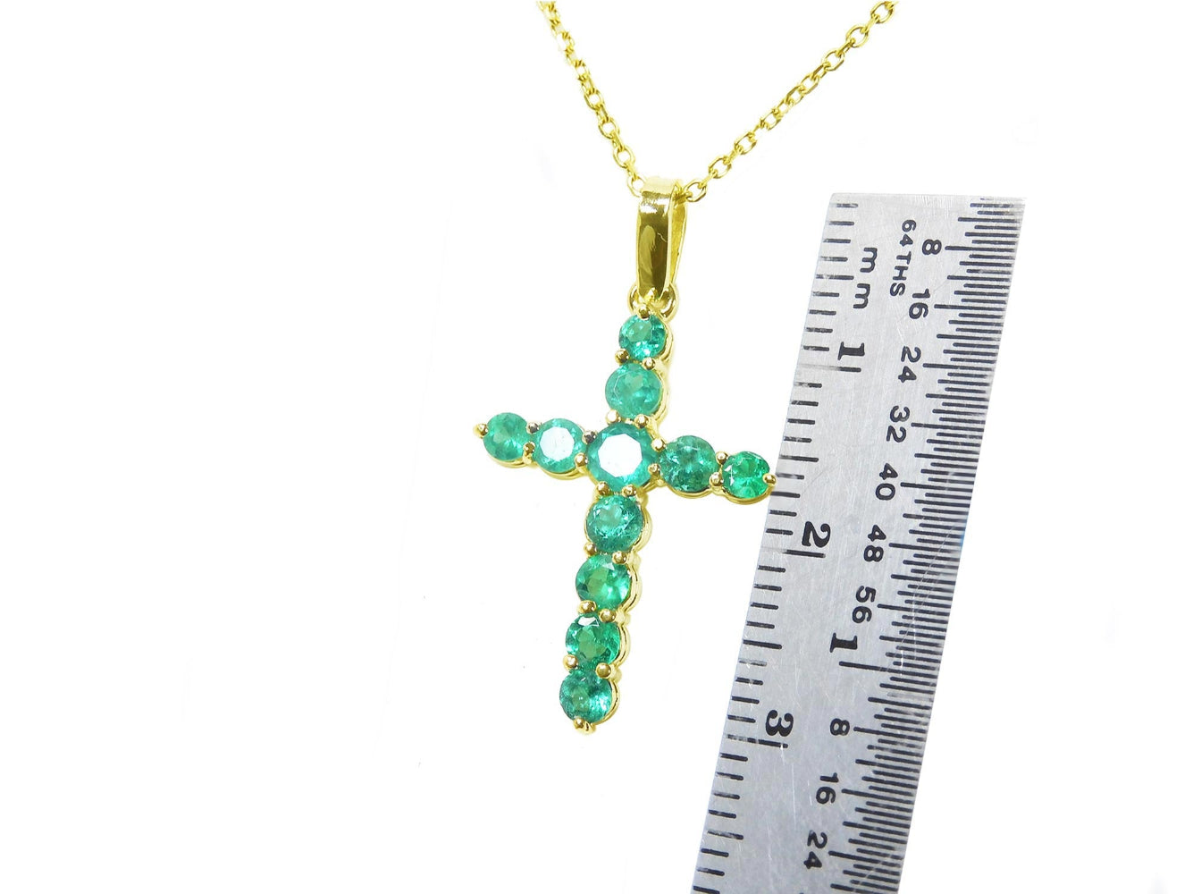 Gold cross pendant with round cut natural colombian emeralds  next to a ruler for scale on a white background.