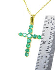 Gold cross pendant with round cut natural colombian emeralds  next to a ruler for scale on a white background.
