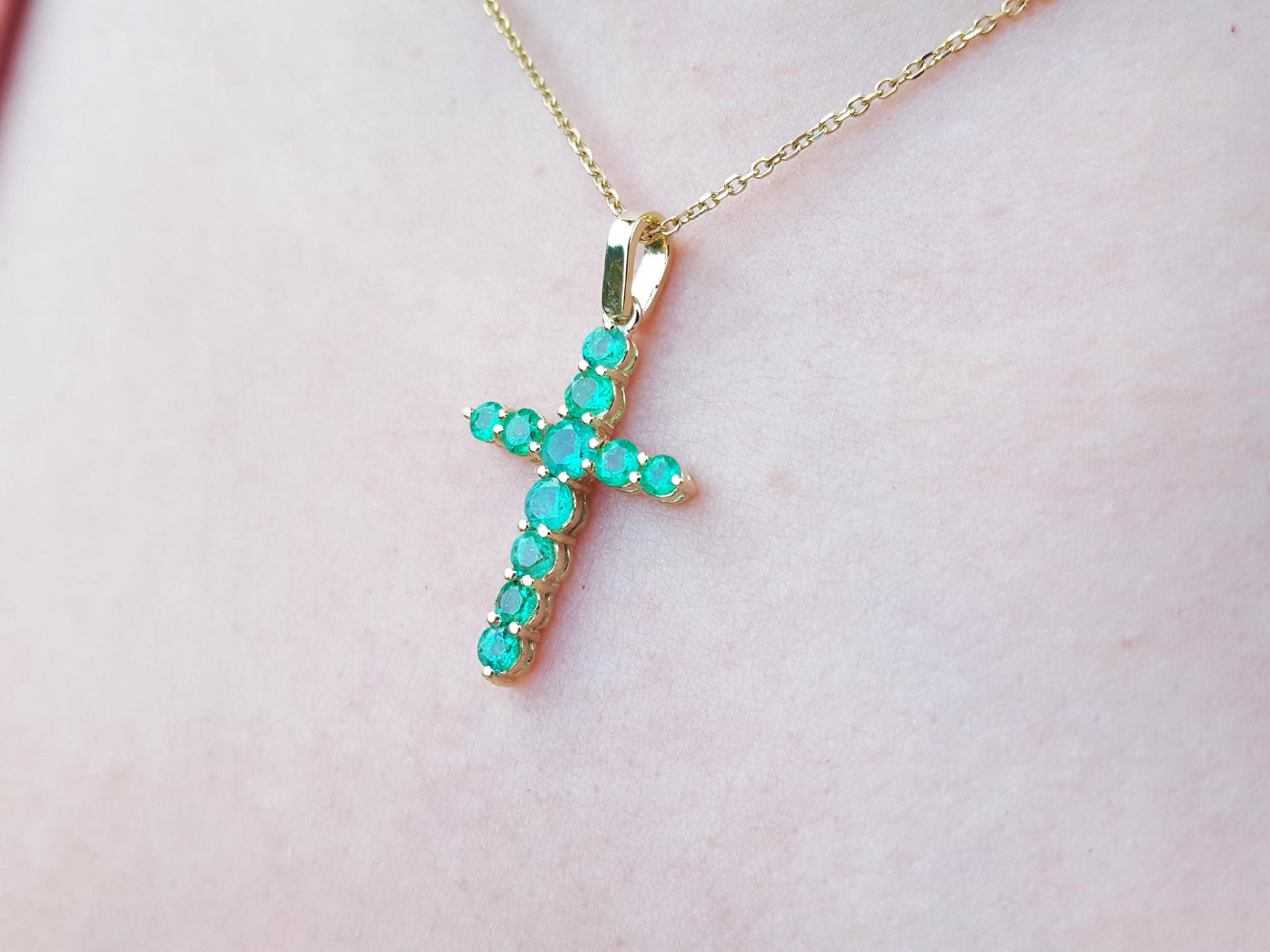 Yellow gold emerald cross pendant on a light pink skin background.