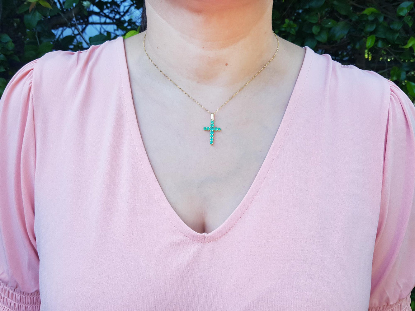 A woman wearing a pink top with a genuine emerald cross necklace against a blurred green background.