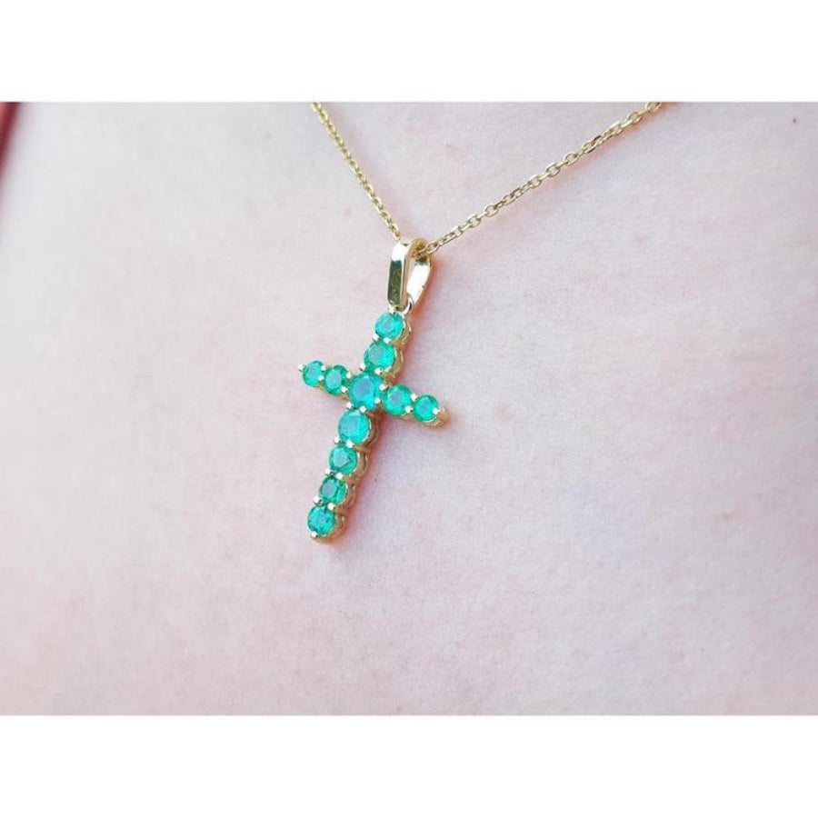 Yellow gold emerald cross pendant on a light pink skin background.