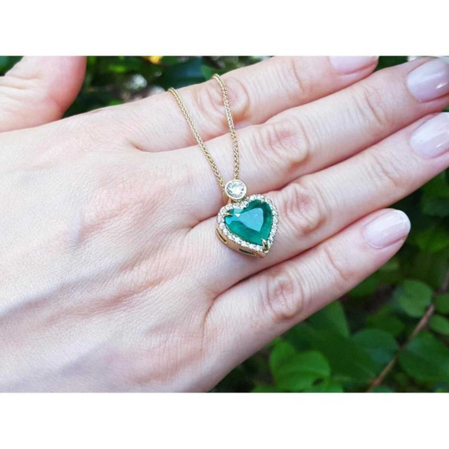 18k gold heart pendant with a bluish-green emerald haloed by diamonds, worn on a hand.
