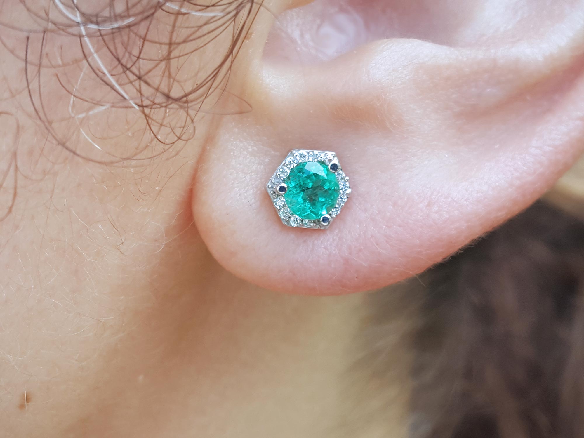 Close-up of an earlobe wearing an hexagonal halo diamond emerald stud earring.