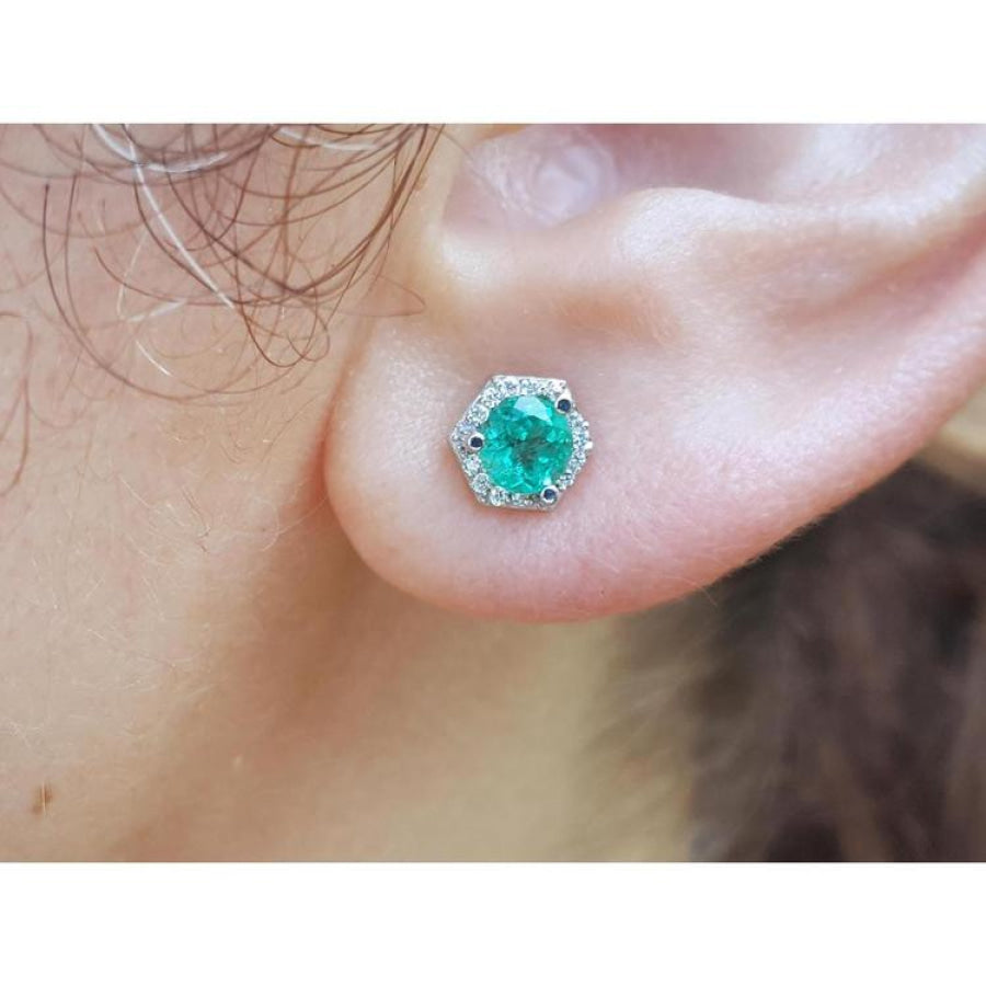 Round cut emerald stud earring in 14K white gold with a hexagonal diamond halo on an ear.