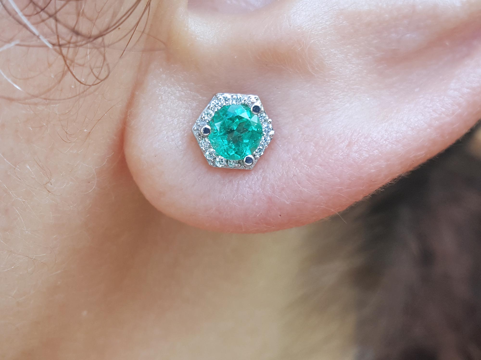 A very close-up of an earlobe dressing an emerald stud earring with hexagonal halo diamond.