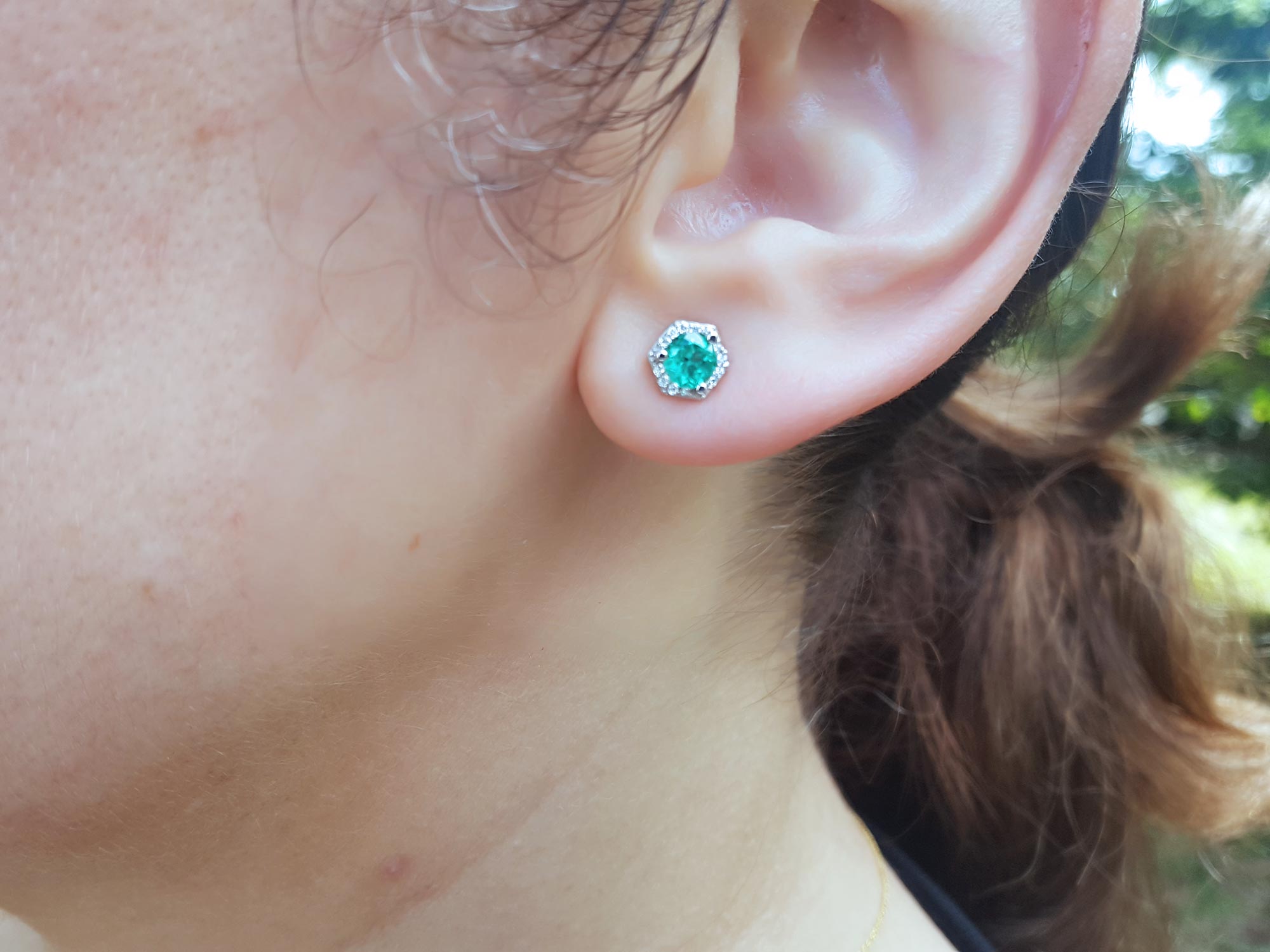A woman&#39;s ear dressing an emerald and diamond stud earring with a blurry background.