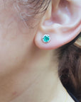 A woman's ear dressing an emerald and diamond stud earring with a blurry background.