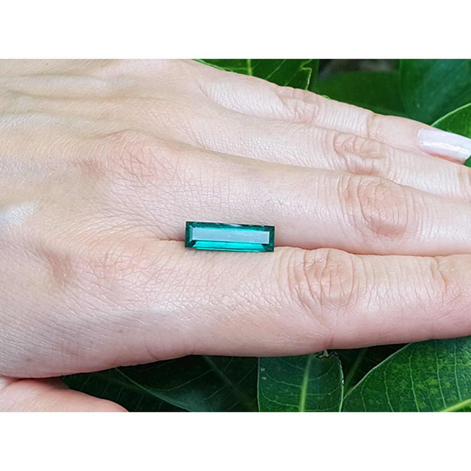3.13 carat baguette cut Colombian emerald, eye clean, sparkling bluish-green, held on a hand with green leaves in the background.