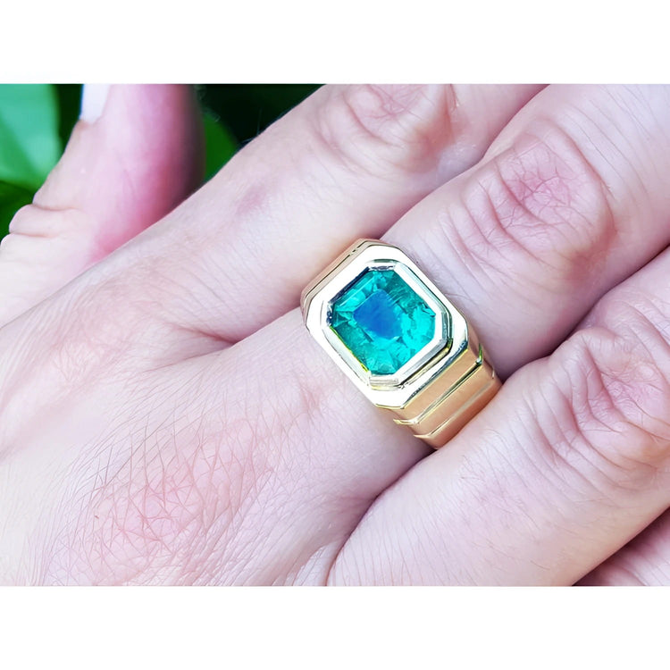 A 18k yellow gold ring for men with a natural colombian emerald on a hand against a blurred natural background.