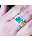 A 18k yellow gold ring for men with a natural colombian emerald on a hand against a blurred natural background.
