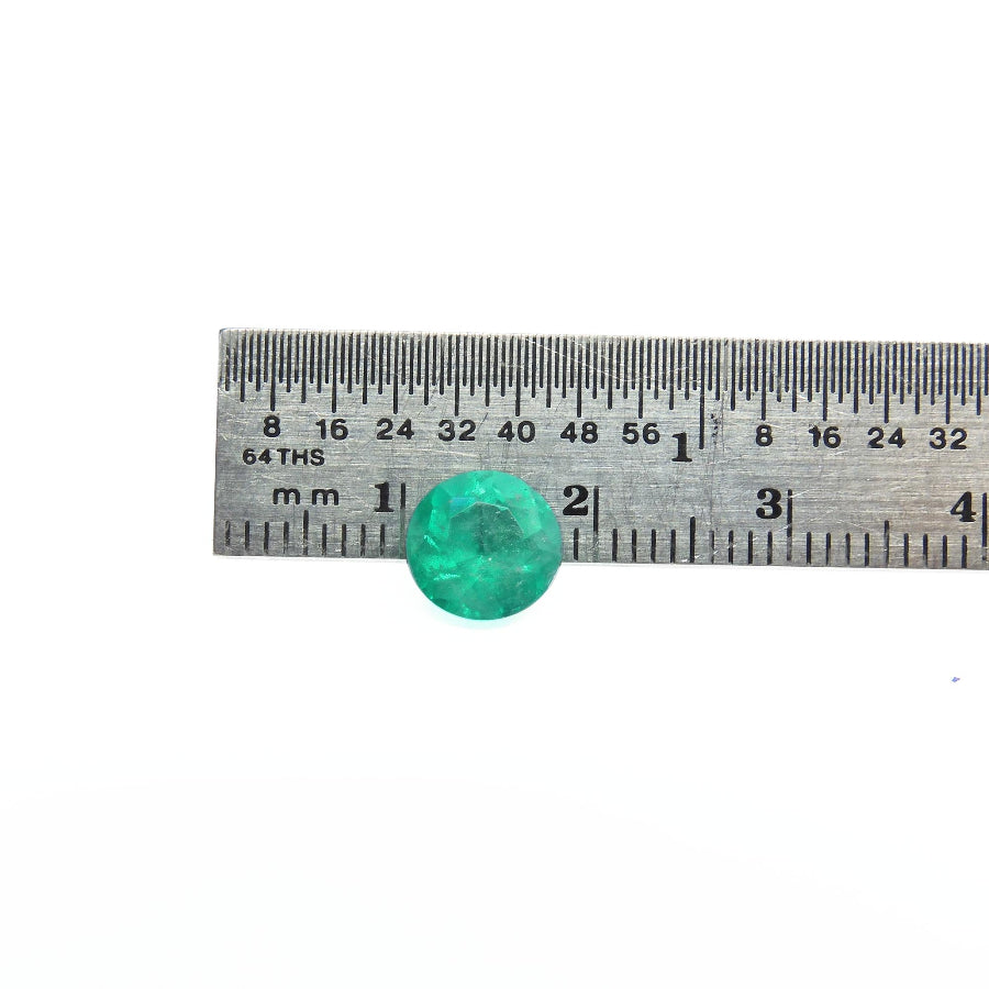 A round cut loose colombian emerald, shown resting on a metal ruler with millimeter markings to indicate its size.