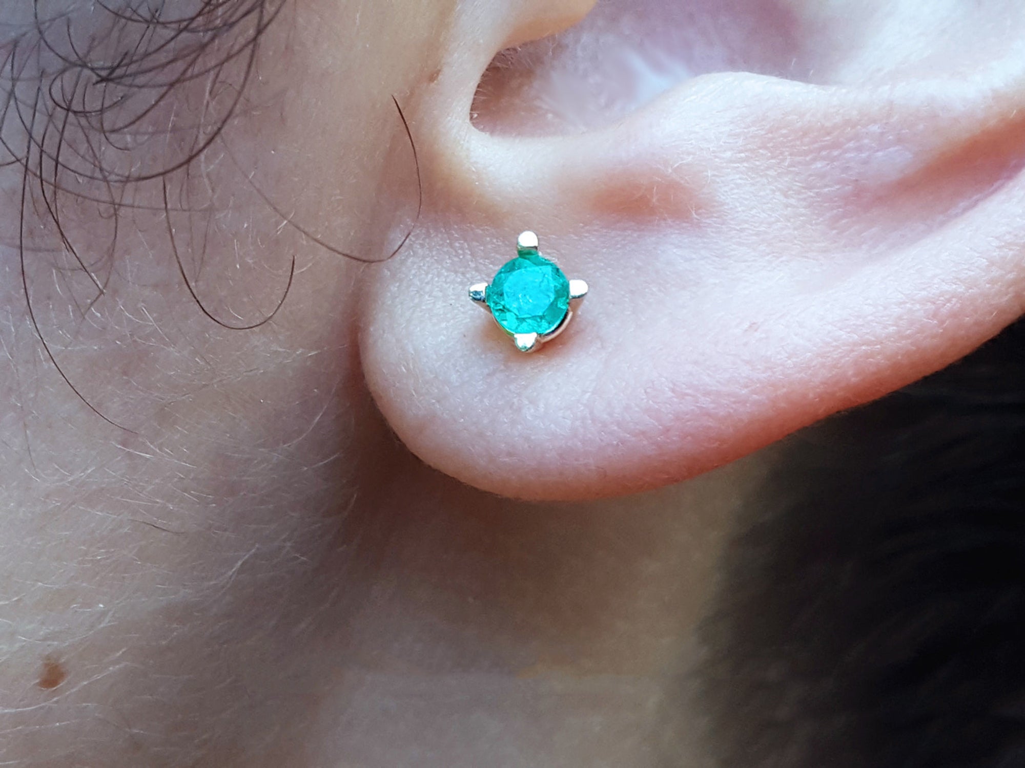 Close-up of an ear wearing a emerald stud earring with a blurred natural background.