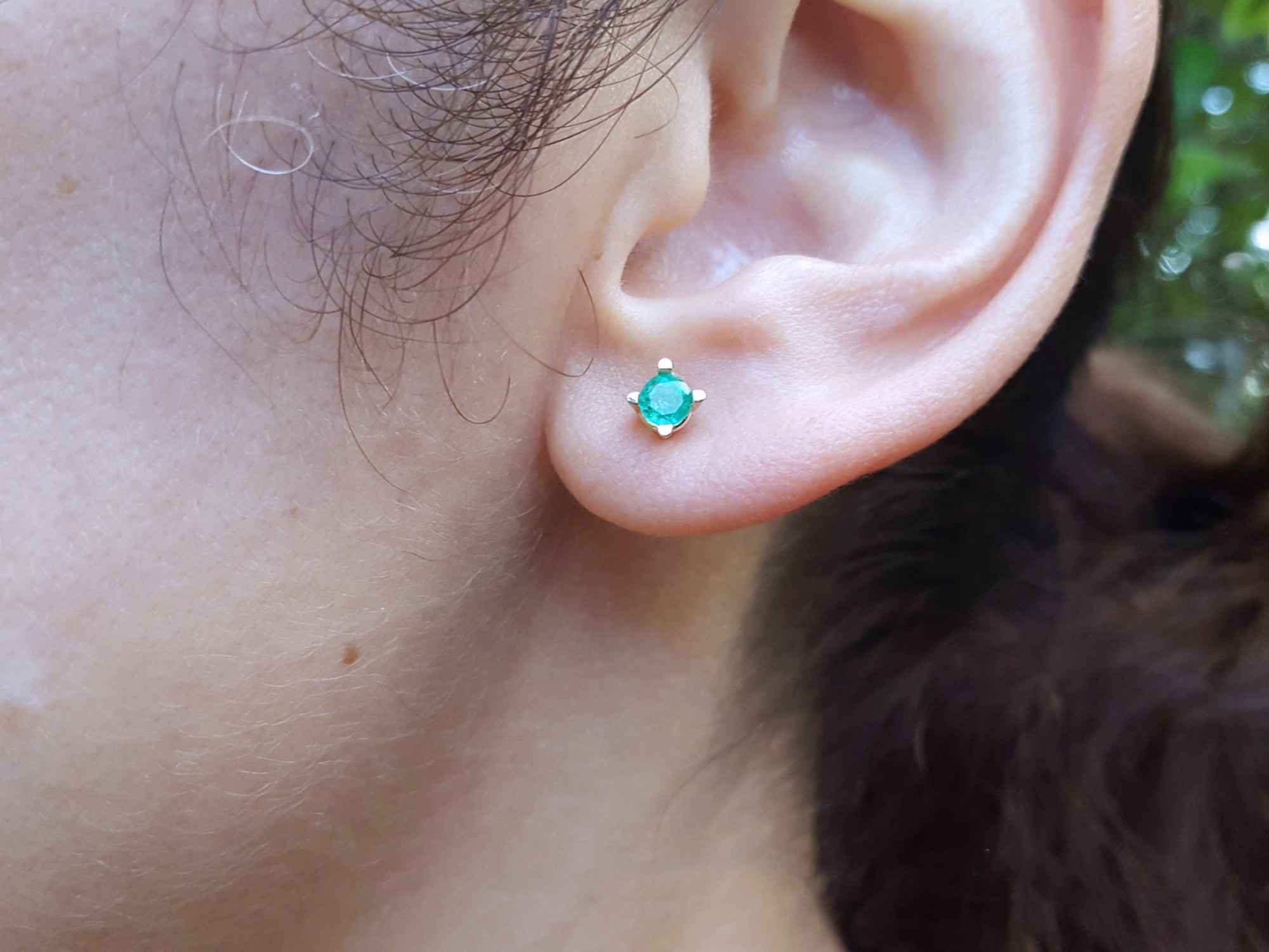 Close-up of an ear wearing a emerald stud earring with a blurred natural background.