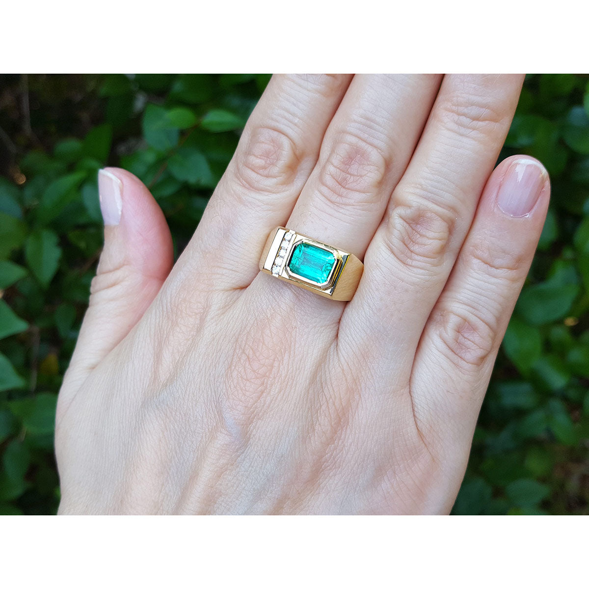 Hand wearing a yellow gold ring with a emerald gemstone against a blurred green background.