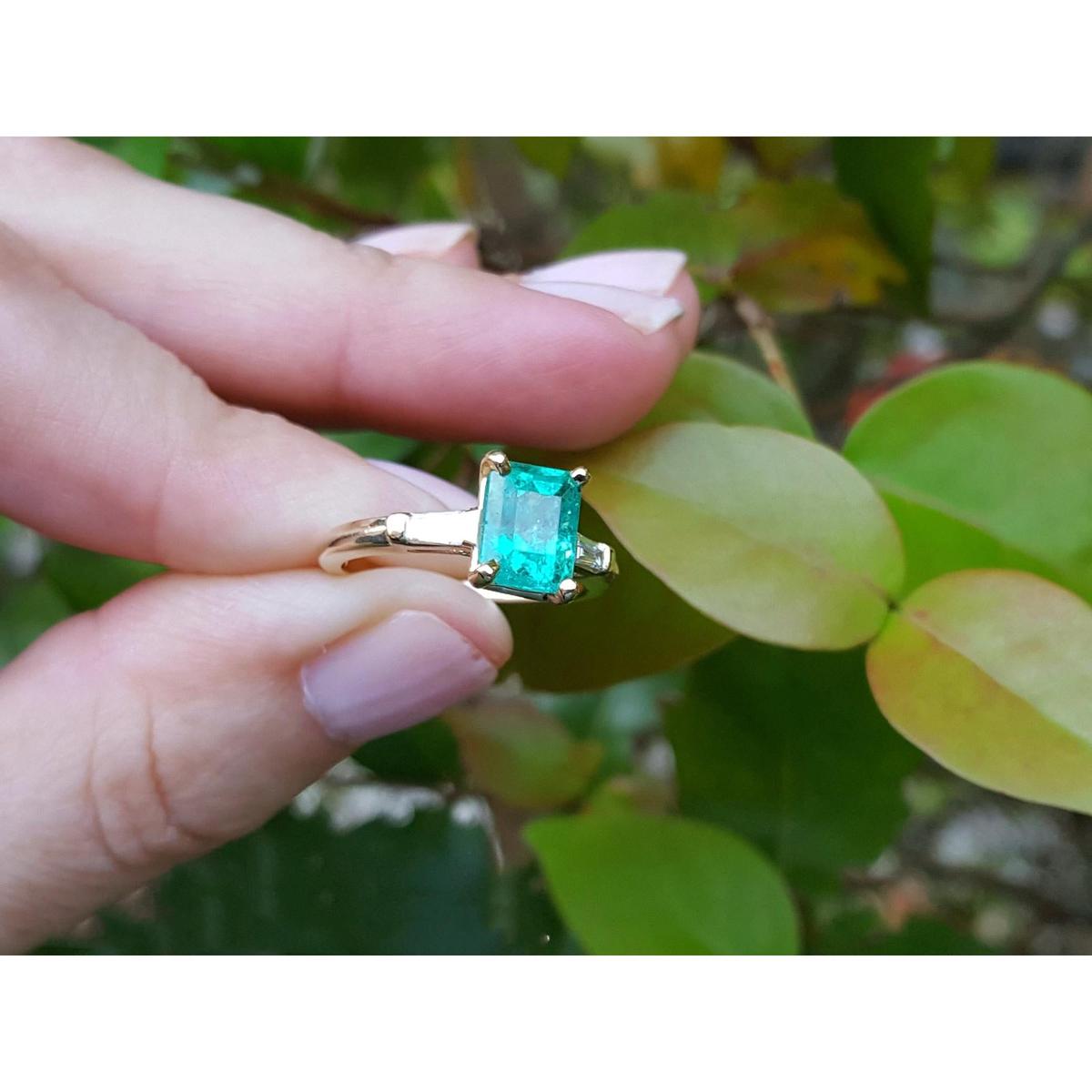Gold ring with a colombian emerald held between fingers against a leafy background.