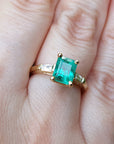 A 14k yellow gold emerald ring with side diamonds on a hand.