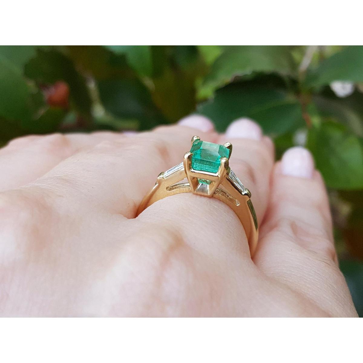 A yellow gold emerald ring with side diamonds on a hand against a blurred green leafy background.