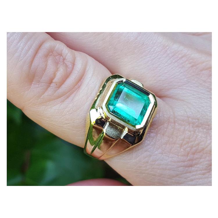 Gold ring with a square natural emerald on a finger against a blurred green background.