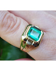 Gold ring with a square natural emerald on a finger against a blurred green background.