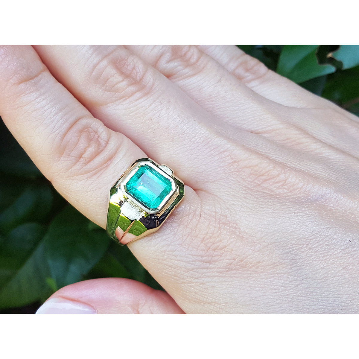 A men&#39;s gold ring with a bezel set natural emerald on a hand against a natural background.