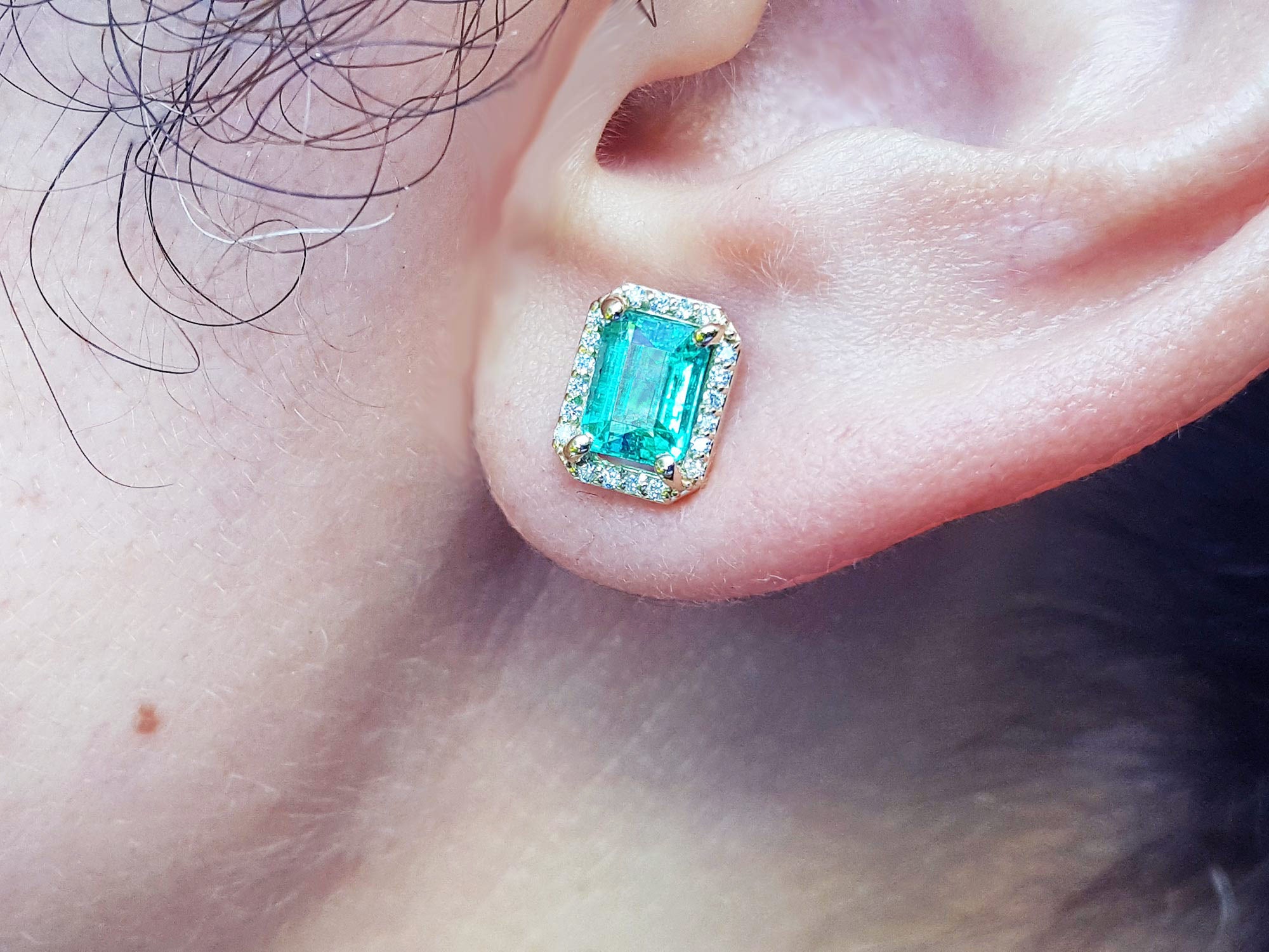 Close-up of an emerald gemstone earring with halo diamond on a person's ear.